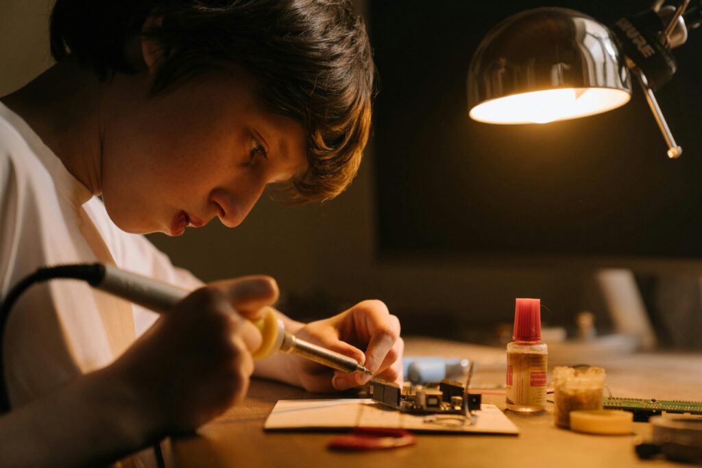 Teenager skillfully soldering a circuit board under a desk lamp, focusing on precision and detail.