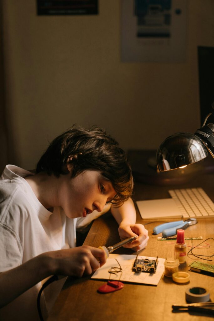 A boy focused intently on repairing a circuit board using a soldering iron at his desk.