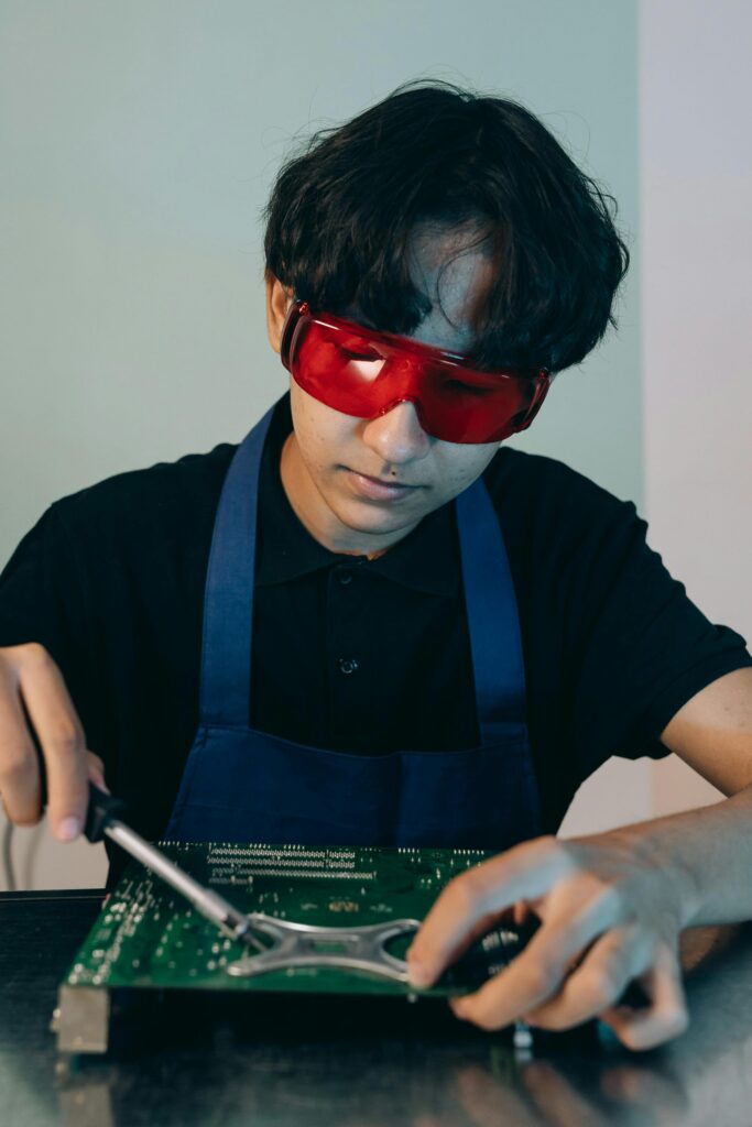 A technician wearing red safety glasses soldering a circuit board indoors.